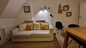 A cozy living room with a sofa, a dining table, and a chair. There is a picture on the wall and a lamp.