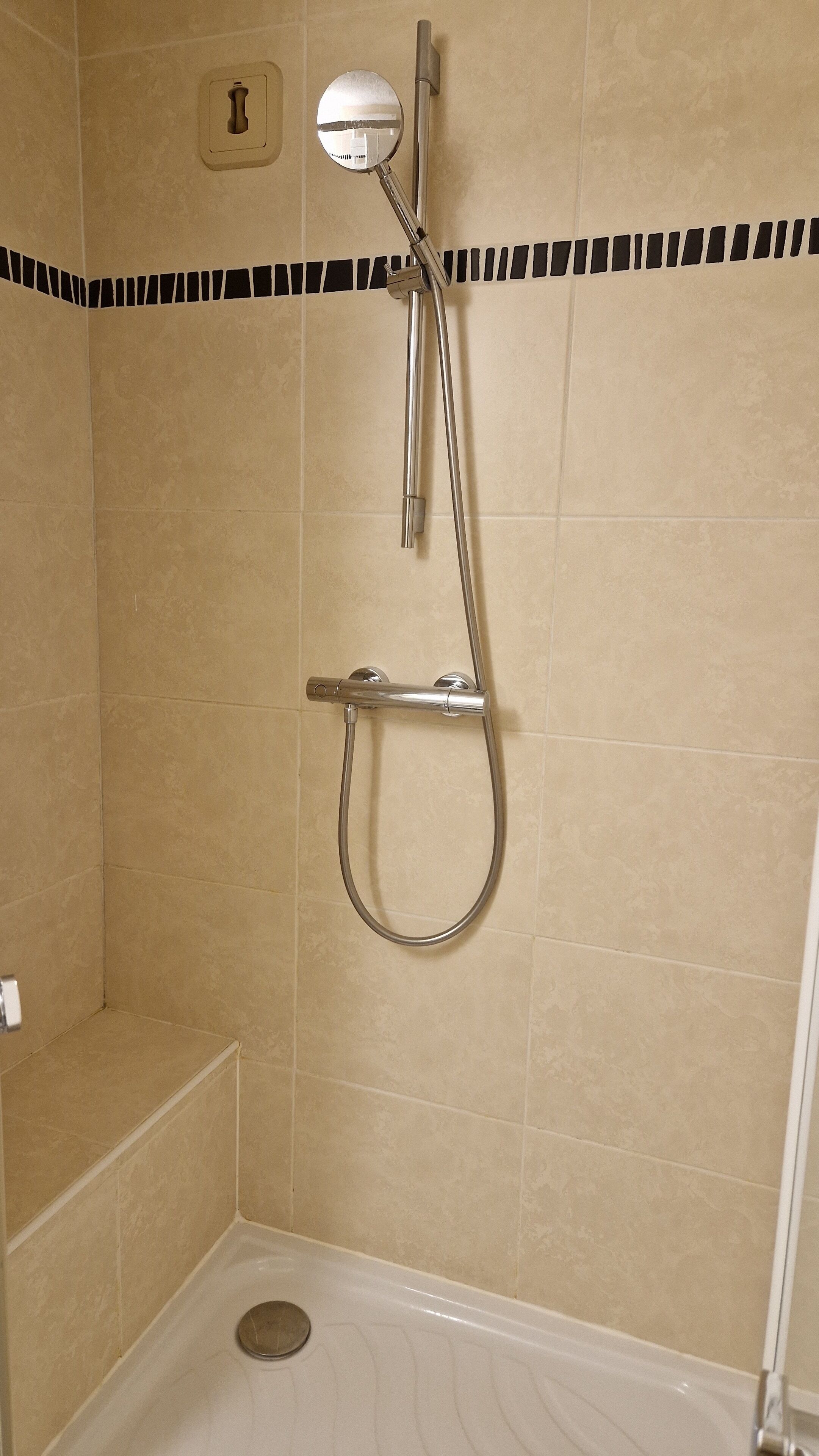 A shower with a handheld showerhead and a wall-mounted faucet, set in a room with beige tiled walls and a black and white border at the top.