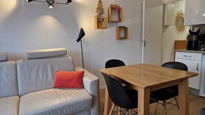 A modern living room with a sofa, a wooden dining table, and chairs. There is a small kitchen area with a coffee maker and a decorative mirror.