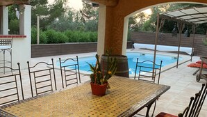 An outdoor patio with a tiled table and chairs, a pool, and a pergola with hanging decorations. - La Campanette in Luberon, private pool. (Cheval Blanc)