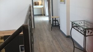Un pasillo con una escalera de madera a la izquierda, una mesa metálica a la derecha y un escritorio con una lámpara al fondo.