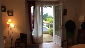 A room with a television, a table, a chair, and a lamp. There is a door leading to an outdoor area with a view of a garden and a building. - F2 on Bordagain Hill. Ciboure (Ciboure)