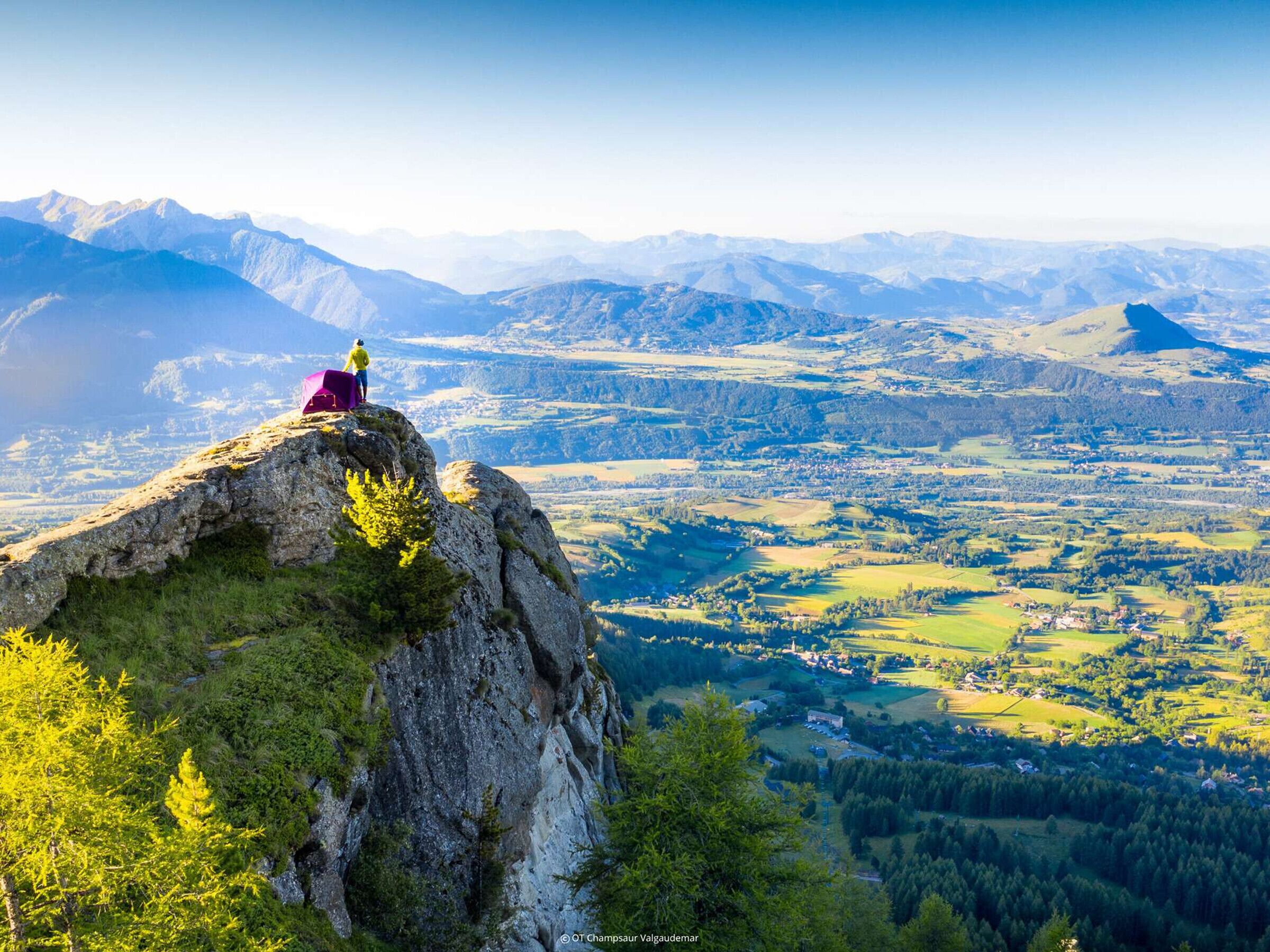 A person standing on a rocky outcrop with a view of a valley and mountains in the background.