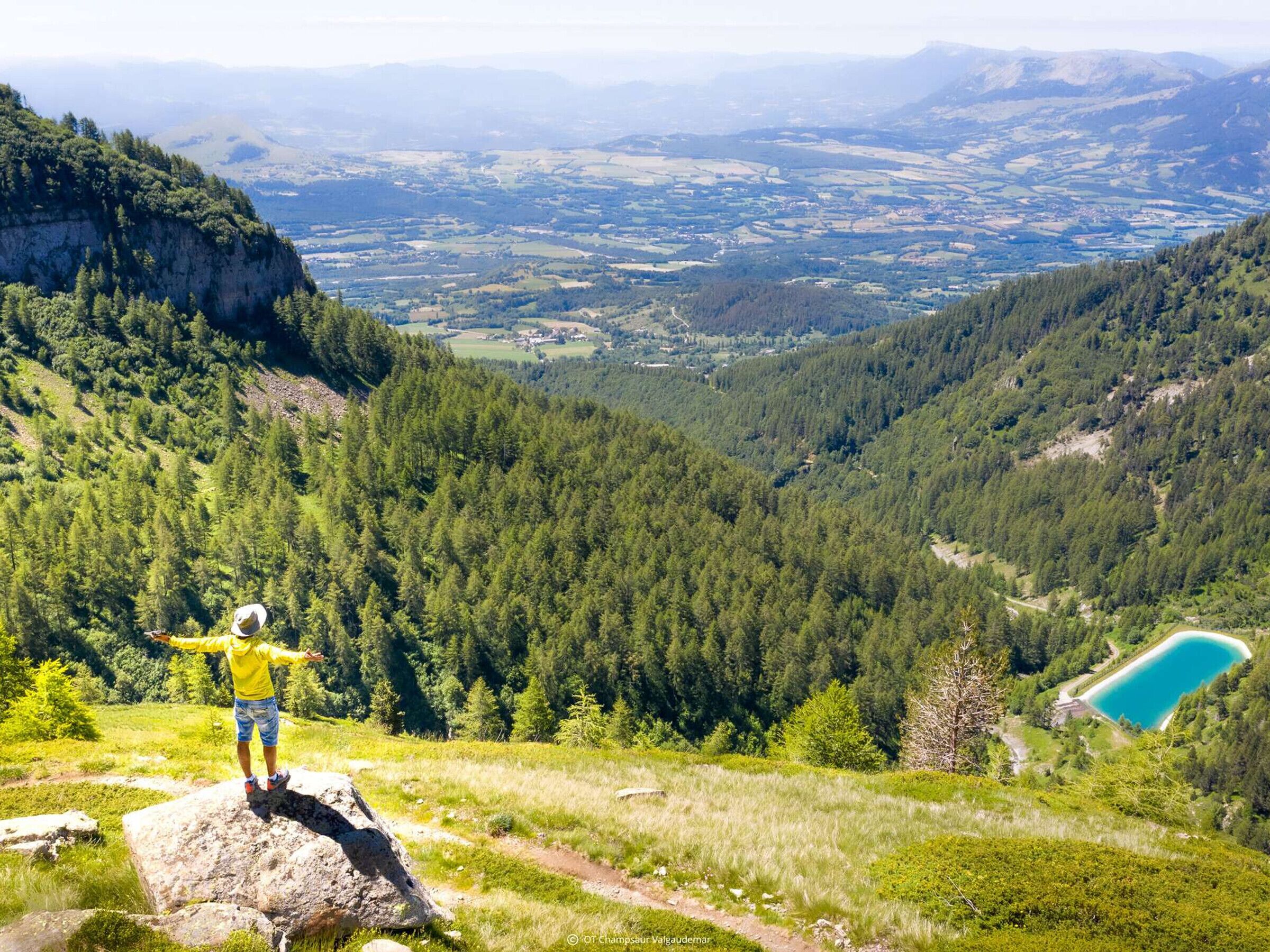 A person standing on a rock with a scenic view of a valley and a lake.