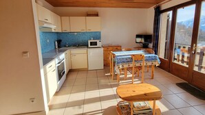 A compact kitchen with a microwave, sink, and wooden cabinets. There is a dining area with a table and chairs, and a small wooden stool. The room has a view of mountains through large windows. - Spacious studio for 4 with balcony in Saint-Michel-de-Chaillol (Saint-Michel-de-Chaillol)