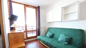Une pièce avec un canapé vert, un bureau en bois avec une lampe, une porte-fenêtre coulissante et un meuble blanc placé au-dessus du canapé.