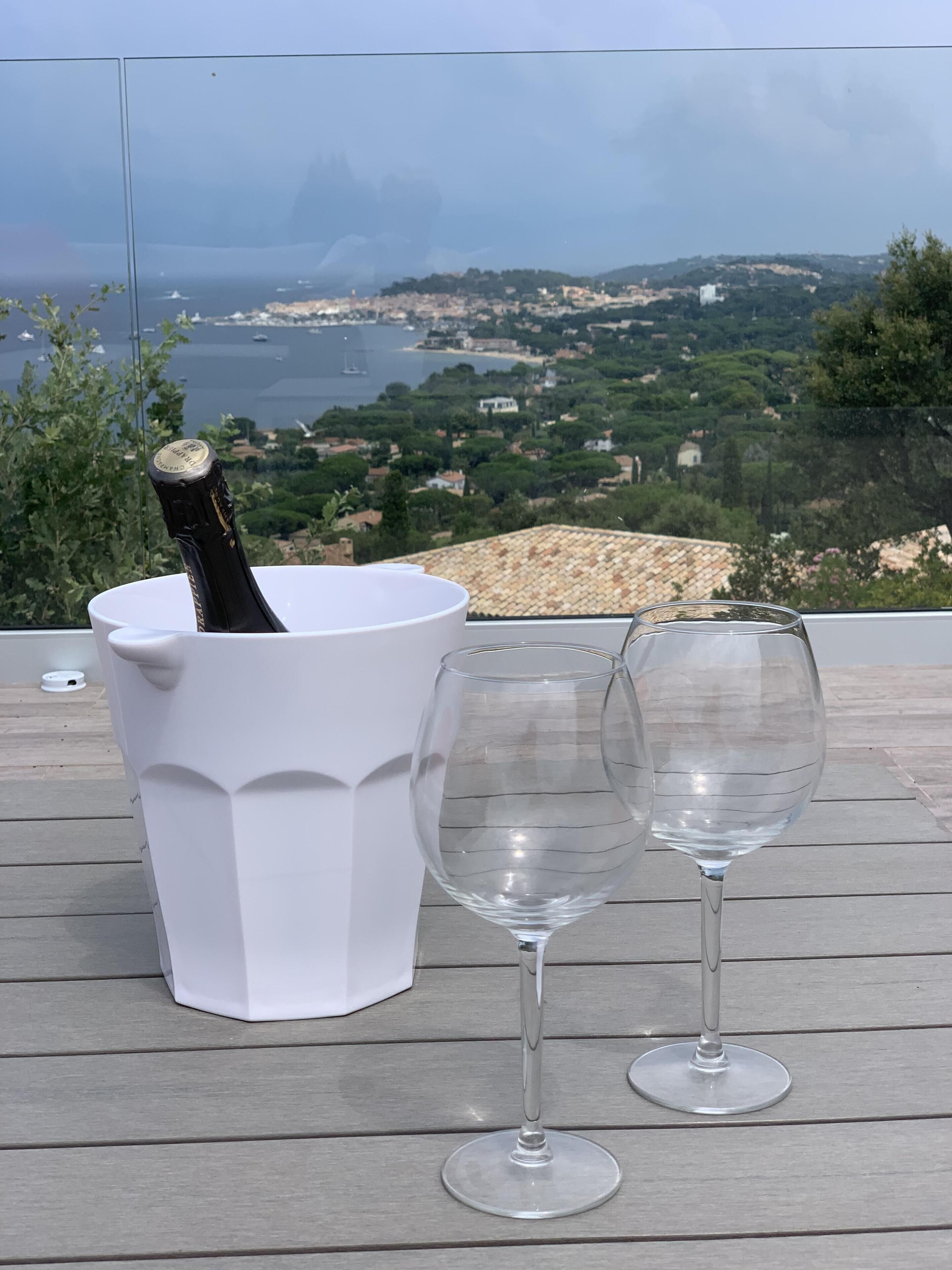 Een witte emmer met een champagnefles en twee lege wijnglazen op een houten terras, met een prachtig uitzicht op een kustplaatsje en glooiende heuvels op de achtergrond.