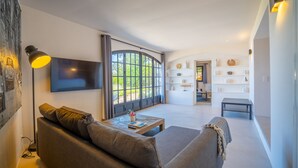 Een moderne woonkamer met een bank, salontafel en een flatscreen-tv. Er is een groot raam met gordijnen, een boekenkast en een kleine tafel.