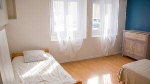 Una habitación con dos camas individuales, un armario de madera y una ventana con cortinas traslúcidas.