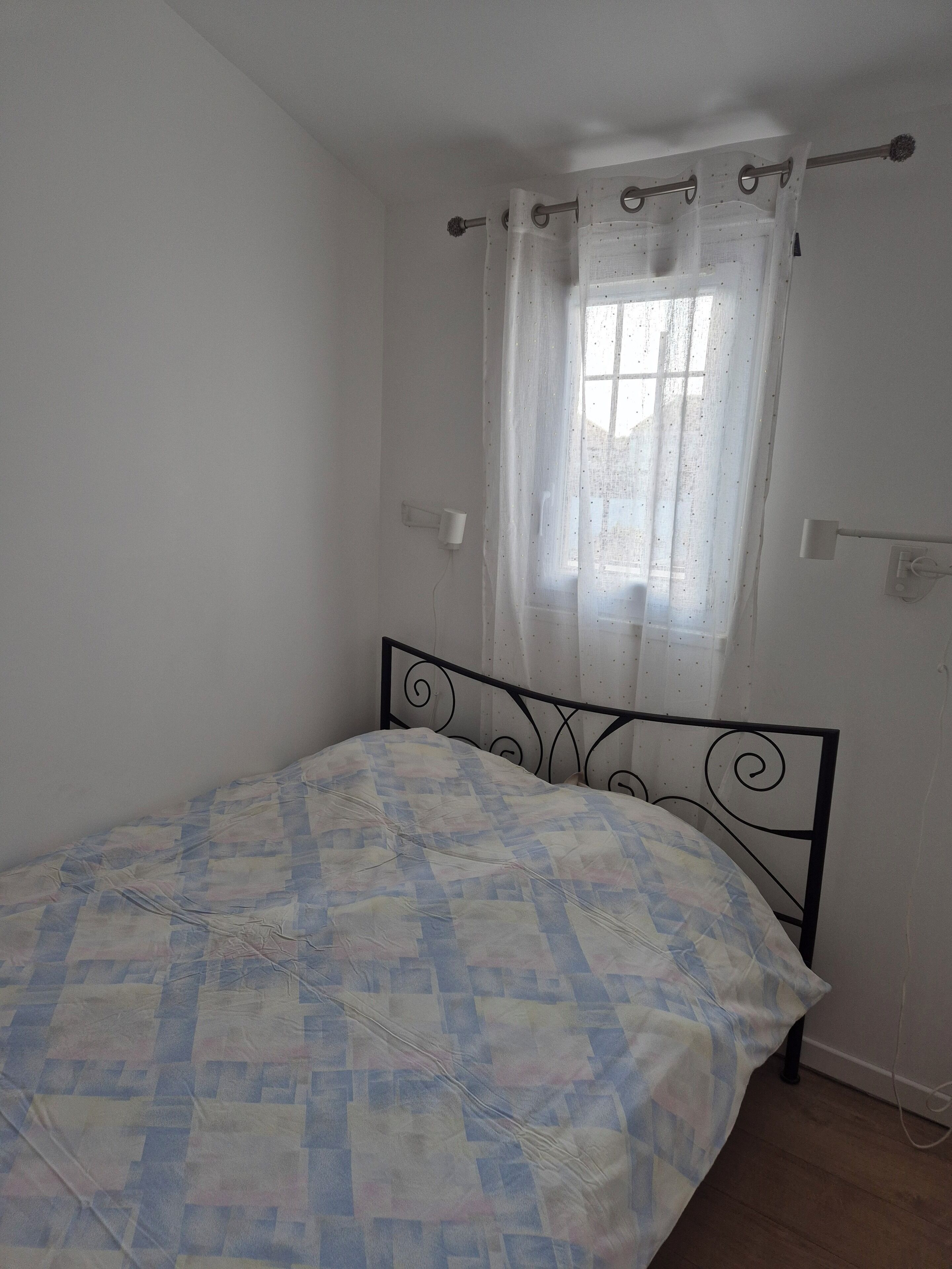 Una habitación pequeña con cama individual, cabecera metálica, ventana con cortinas traslúcidas y pared blanca lisa.