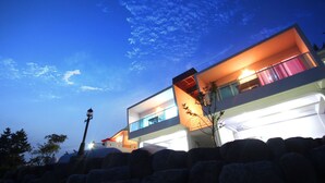 Un edificio moderno de dos pisos con un balcón y un interior iluminado, sobre un cielo crepuscular.