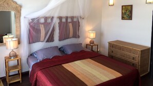 Un dormitorio con cama, una mesita de noche con lámpara, un tocador, un espejo, un aparador y un cuadro en la pared.