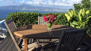 Una mesa de madera con un jarrón de flores, rodeada de sillas, con vista al mar, montañas y un paisaje urbano.
