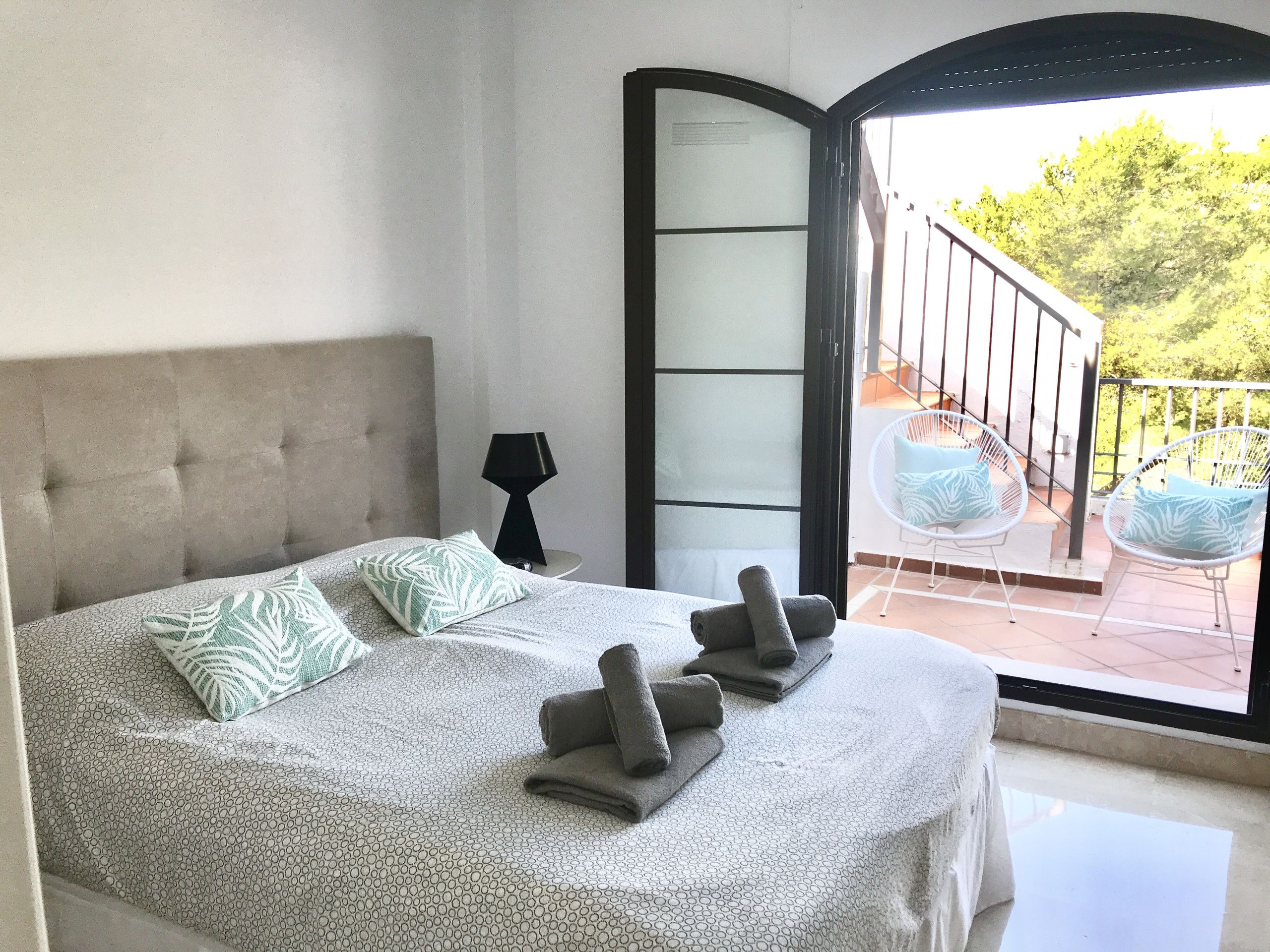 A bedroom with a bed, pillows, a nightstand, a lamp, and a view of an outdoor balcony with chairs.