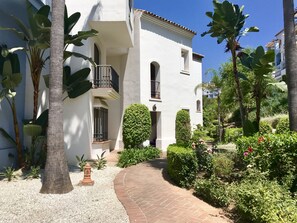 A white building with a brick pathway leading to a gated entrance, surrounded by lush greenery and palm trees.