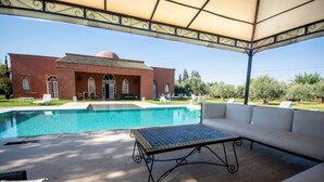 Zona de piscina con área de descanso cubierta, mesa de billar y un edificio con ventanas arqueadas al fondo.
