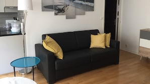 Sala de estar com sofá preto, duas almofadas amarelas, mesa de centro de vidro, abajur branco e um quadro na parede com uma cena de praia.