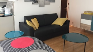 Uma sala de estar com um sofá preto, uma mesa de centro com toalha de mesa listrada e uma poltrona azul, além de outra mesa de centro verde-água.