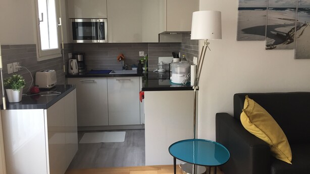 Uma cozinha compacta com eletrodomésticos modernos, um pequeno espaço de refeições e uma sala de estar com sofá e luminária.