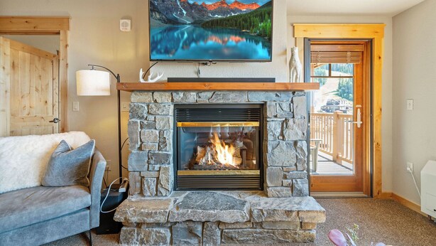 A living room with a stone fireplace, a mounted TV displaying a mountain scene, a wooden door leading outside, and a cozy gray sofa.