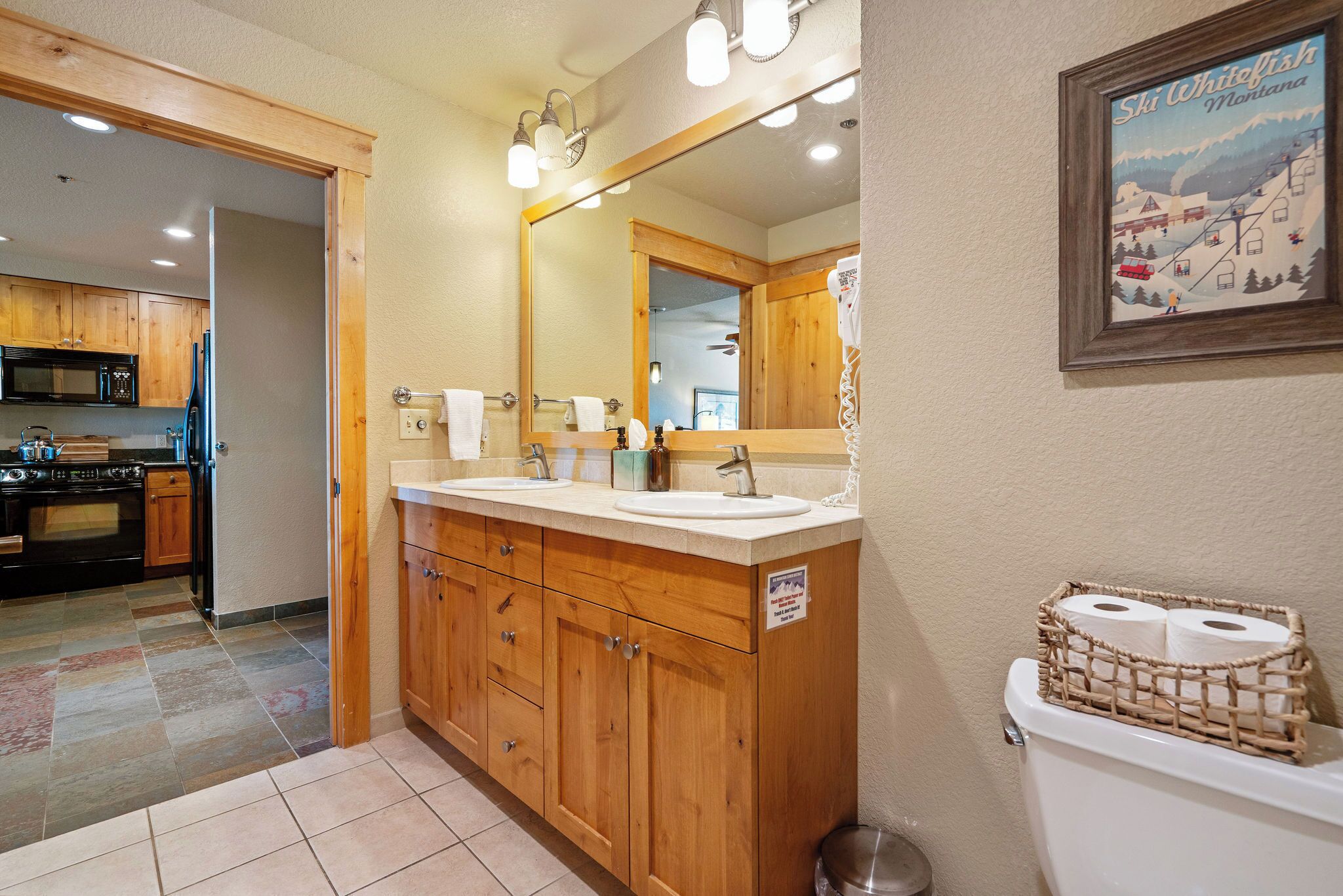 A bathroom with wooden cabinets, a double sink, a large mirror, and a toilet with toilet paper.