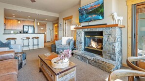 A modern living room with a stone fireplace, a mounted TV, and a kitchen in the background.