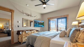 A bedroom with a large bed, a ceiling fan, a mounted TV, and a window with a view.