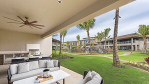 Un espace de vie extérieur moderne comprenant un canapé, une table à manger et un ventilateur de plafond. Il offre une vue sur une pelouse bien entretenue et un immeuble doté de nombreux balcons.