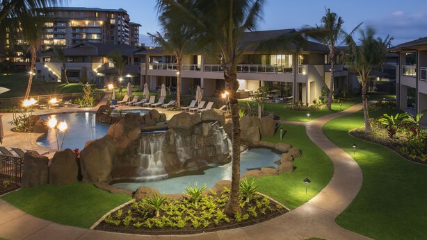 Un espace piscine aménagé dans un complexe hôtelier, avec une cascade, des chaises longues et un chemin bien éclairé.