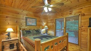 Quarto de cabana de madeira com uma cama, duas mesas de cabeceira, um ventilador de teto, um mapa na parede e uma janela com persianas.