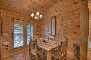 A wooden cabin interior with a dining table, chairs, a chandelier, and a window with blinds. - Ole Bear Paw Cabin - Cherry Log (Cherry Log)