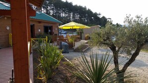 A wooden building with a green roof, outdoor seating with yellow umbrellas, and a gravel area with plants and trees. - St Maurice - Plein Air Chalet C1 (Dieulefit)