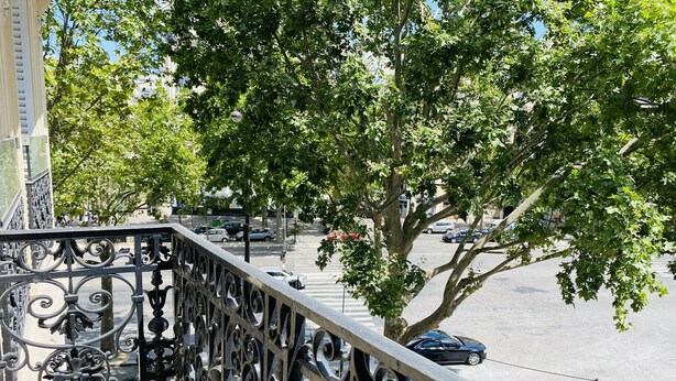 A balcony with ornate metal railings overlooking a street with cars and trees.
