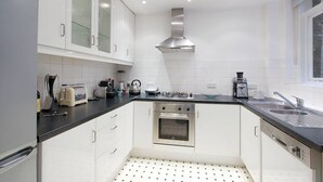 A modern kitchen with white cabinets, a stainless steel range hood, a built-in oven, a black countertop, and a window providing natural light. - Spacious, baby-ready 2br 1st floor in Chelsea (london)