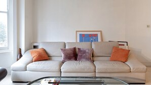 A living room with a beige sofa, two orange and two purple cushions, a glass coffee table, and a framed artwork on the wall. - Spacious, baby-ready 2br 1st floor in Chelsea (london)