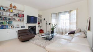 A modern living room with a sofa, armchair, fireplace, bookshelf, and a flat-screen TV. - Spacious, baby-ready 2br 1st floor in Chelsea (london)
