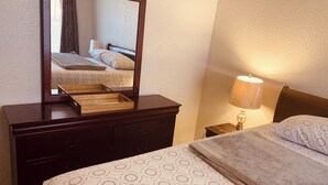 A hotel room with a bed, a wooden dresser, a mirror, a bedside lamp, and a window with curtains. - DENIS TEMPORARY HOUSING 3 BEDROOMS APT- D (Killeen)
