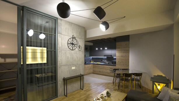 Un salon moderne doté d’un plancher en bois, d’une cloison en verre, d’un coin repas avec une table et des chaises, et d’une cuisine avec un comptoir noir et des éléments en bois.