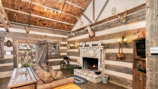 Interior rústico de una cabaña de troncos con chimenea de piedra, sofá de cuero, mesa de madera y zona de comedor.
