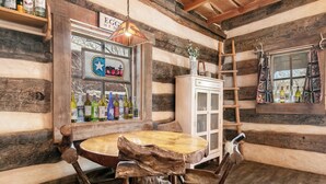 Un interior rústico de madera con una mesa redonda y sillas, un armario con botellas y una escalera apoyada en la pared.