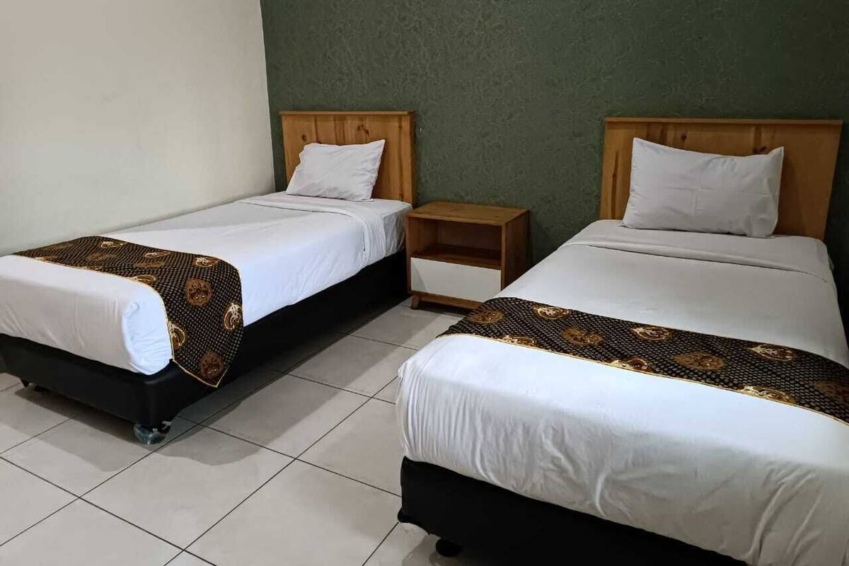 A hotel room with two single beds, each with a wooden headboard and white bedding. There is a small wooden nightstand between the beds.