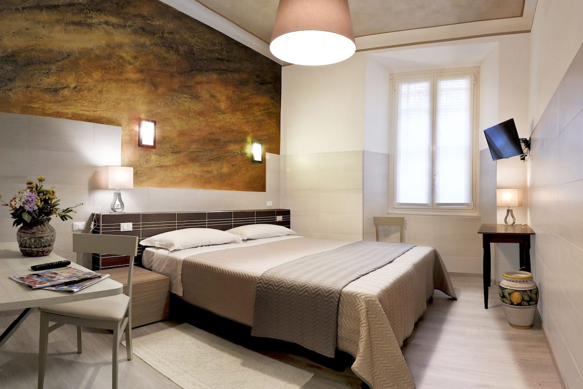 A modern bedroom with a bed, bedside tables, a desk, a chair, a lamp, a TV, and a decorative vase.