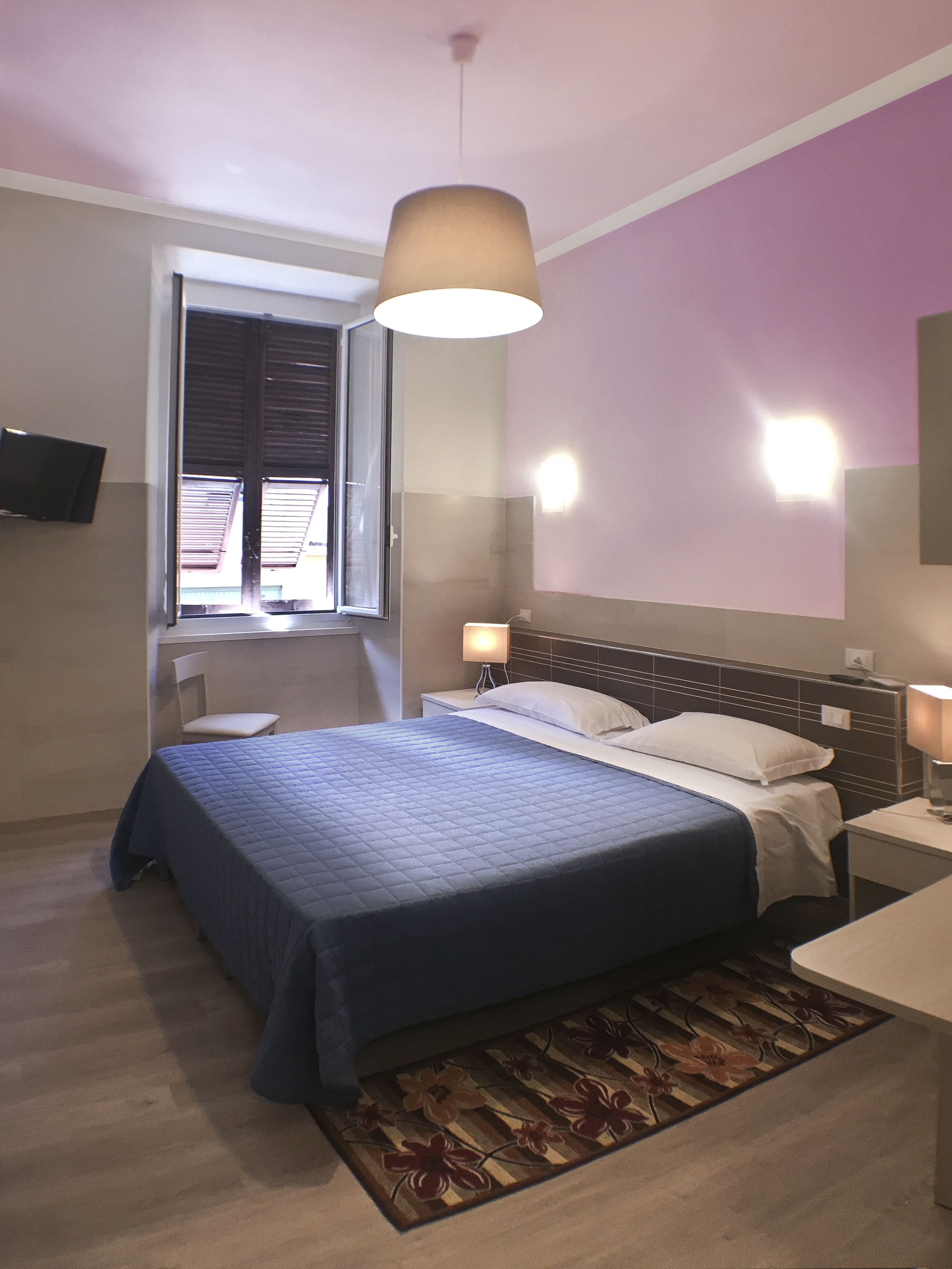 Quarto com cama, mesas de cabeceira, luminária de apoio, janela com persianas, televisão na parede e luz no teto.
