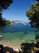 Una spiaggia costiera con acque limpide, persone che nuotano e prendono il sole, e barche all'orizzonte.