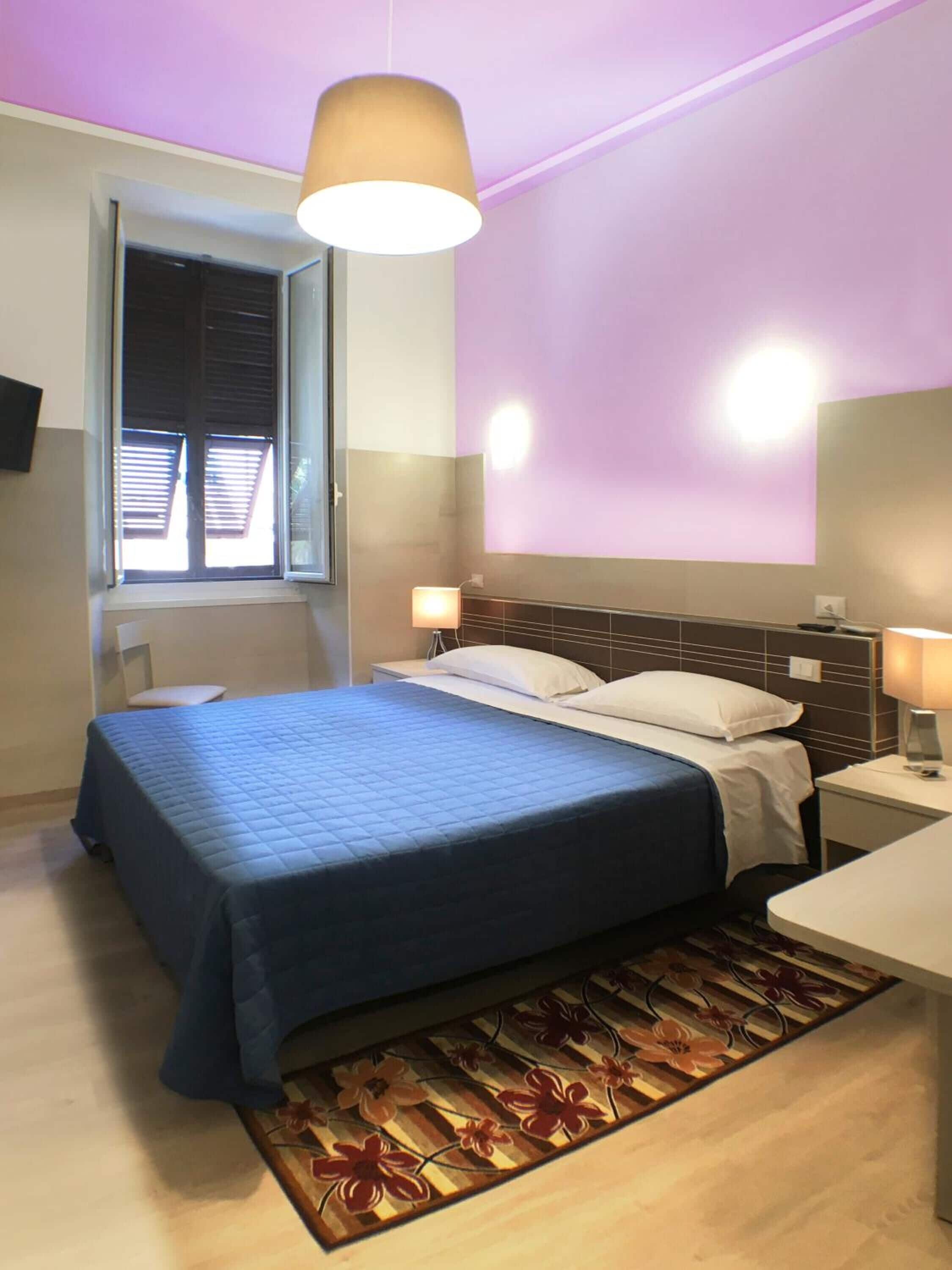 A hotel room with a bed, bedside lamps, a window with blinds, and a wall-mounted light fixture.