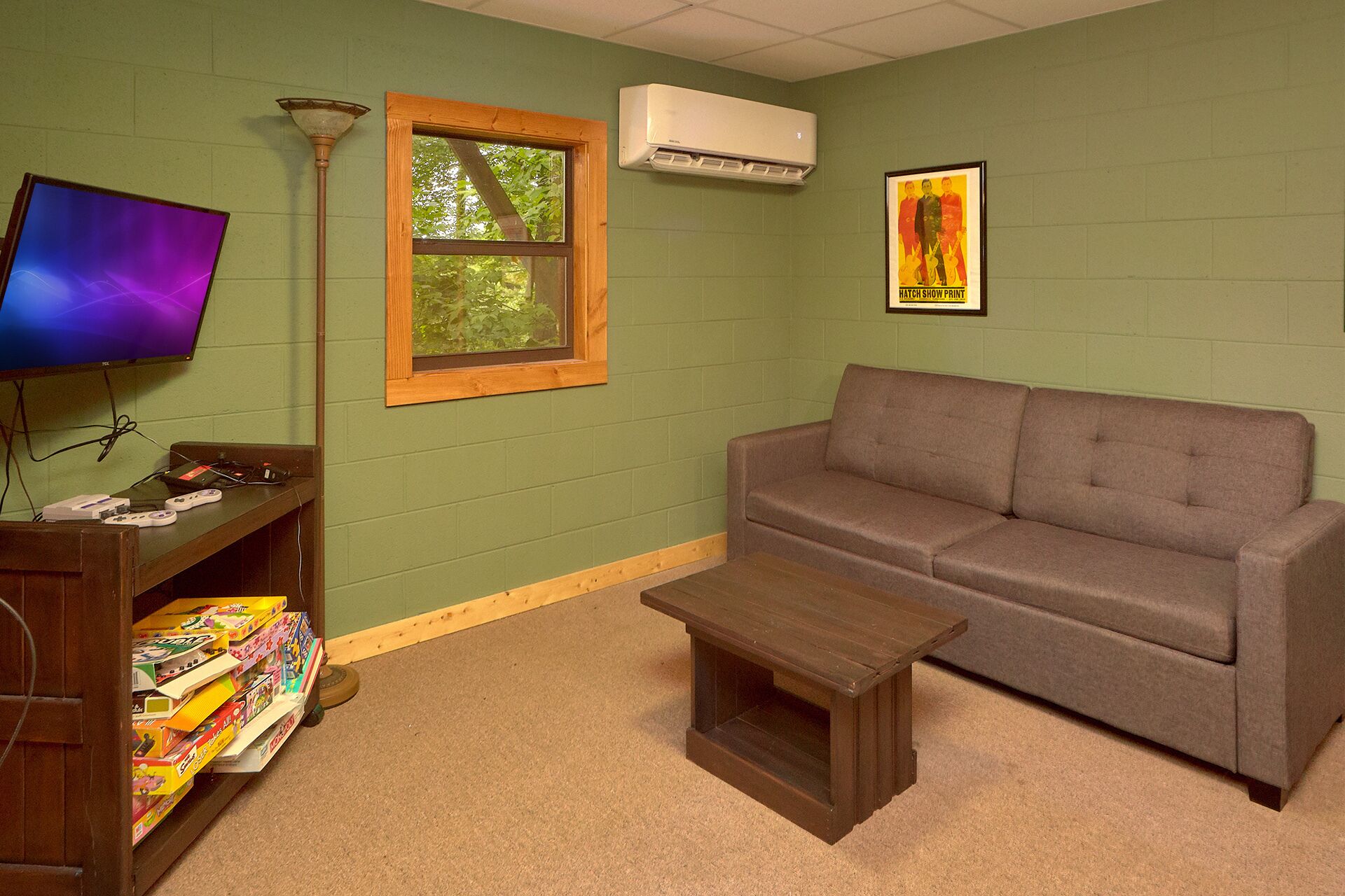 Una habitación con pared verde, un sofá gris, una mesa de centro de madera, un televisor sobre una estantería y un aire acondicionado.