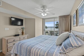Une chambre avec un lit, un ventilateur de plafond, une télévision, une commode, un vase de fleurs et un balcon offrant une vue sur les palmiers.