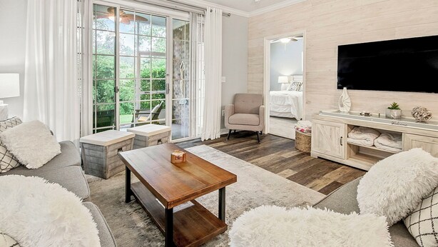 Un salon moderne avec une table basse en bois, des canapés blancs moelleux, un téléviseur fixé au mur et une porte-fenêtre coulissante donnant sur un jardin.