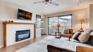 A living room with a fireplace, a television, a ceiling fan, a sofa, and a view of a snowy landscape through large windows.