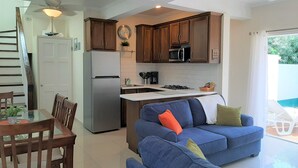 A modern kitchen with dark wooden cabinets, a stainless steel refrigerator, and a built-in microwave. There is a dining area with a glass table and wooden chairs. A blue sofa with cushions is placed in the living area, and a ceiling fan is visible. - Sugar Fields Hoilday Home with Private Pool. (Valley Church)
